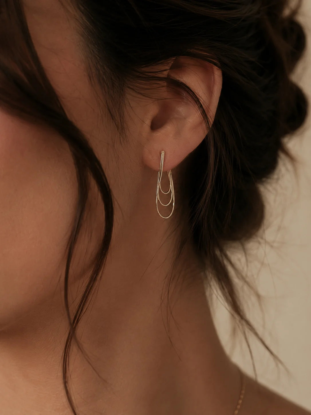 A close-up of a model's ear wearing a delicate gold earring with three stacked, curved oval loops.