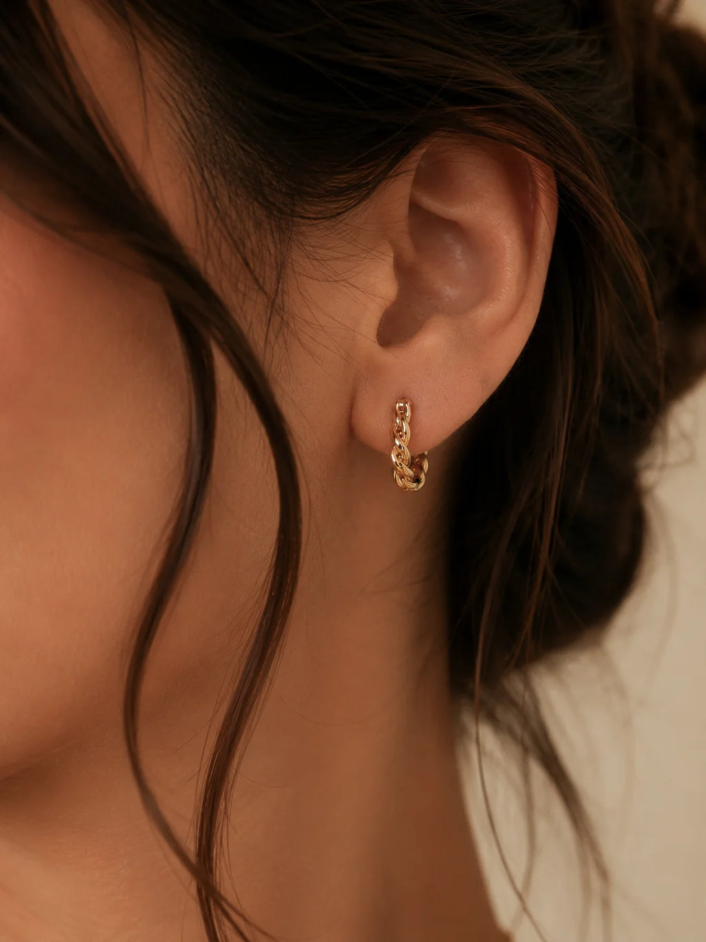 A close-up of a person's ear wearing a small gold huggie hoop earring with tiny clear stones, against a soft beige background.