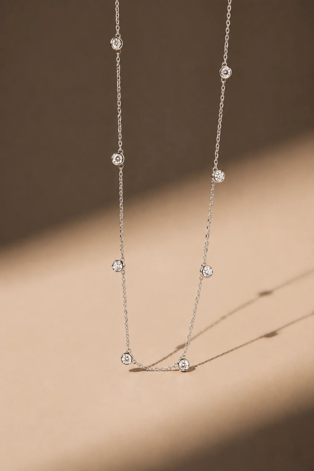 A delicate silver chain necklace with multiple small, round bezel-set clear stones, photographed hanging against a warm beige background with soft shadows.