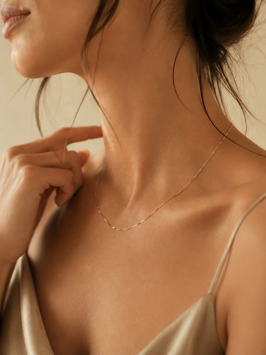 A close-up lifestyle shot of a model wearing a delicate, thin gold chain necklace with a small pavé-style center detail. The model is wearing a champagne-colored satin camisole against a warm beige background.