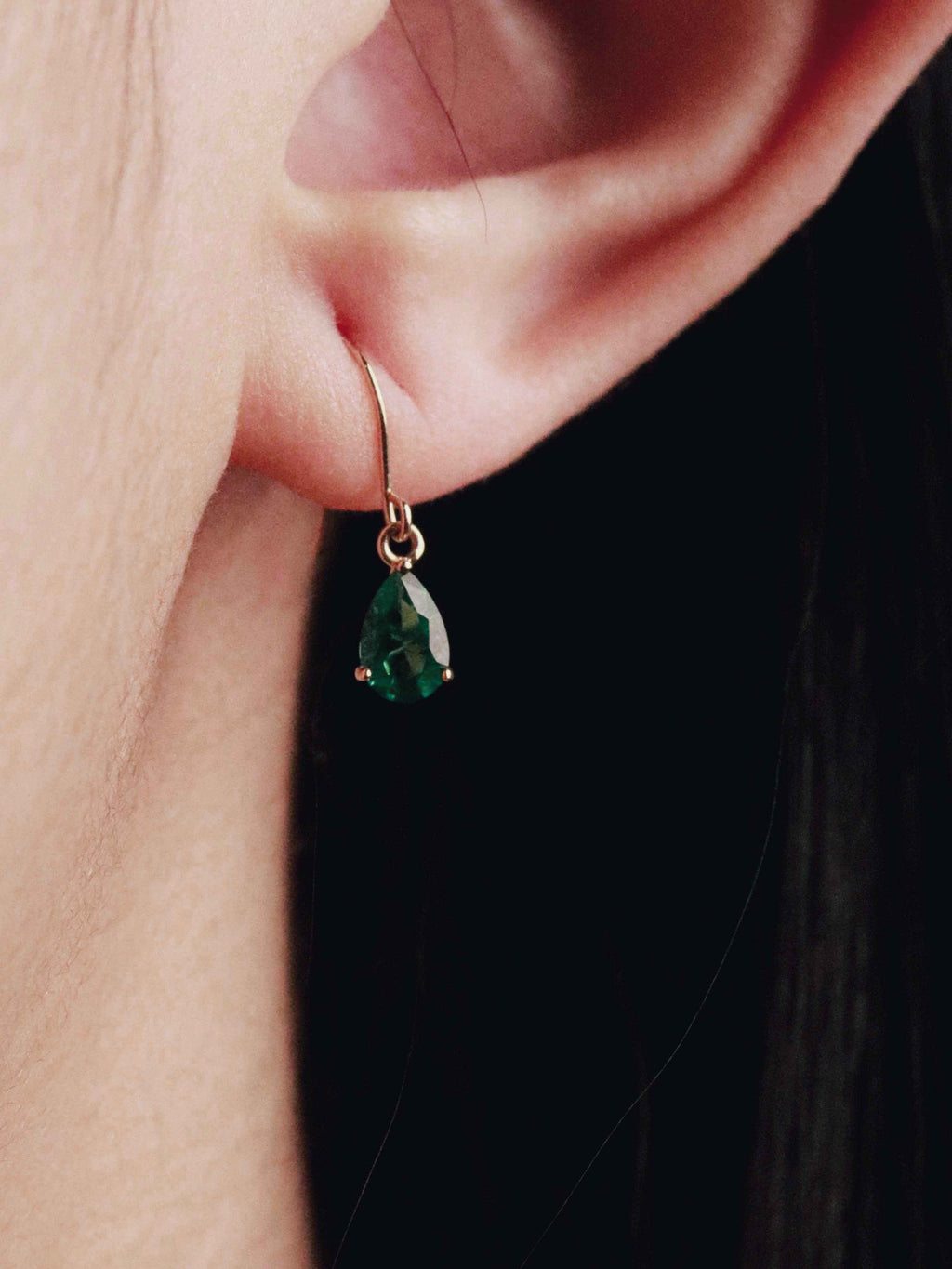 Close-up of an ear wearing a gold earring with a green gemstone against a dark background.