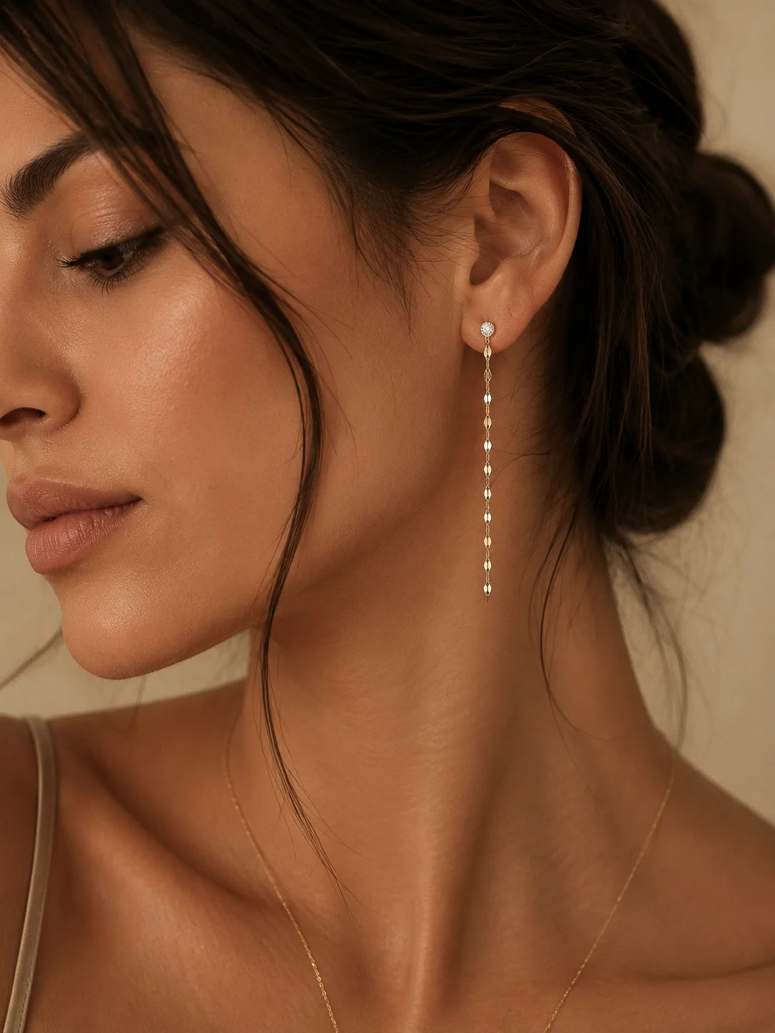 A close-up of a woman wearing a delicate gold dangle earring with small clear stones, shown against a warm beige background.