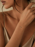 A close-up lifestyle shot of a model wearing delicate gold chain bracelets on her wrist, featuring small green, black, and white gemstone accents, paired with a satin champagne slip dress.