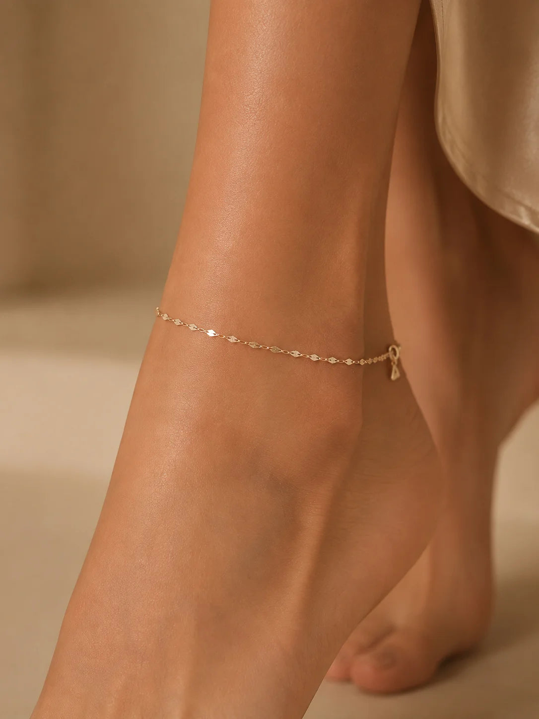 A delicate gold chain anklet with a small dangling charm worn on a person's ankle, shown in a warm, softly lit close-up shot.
