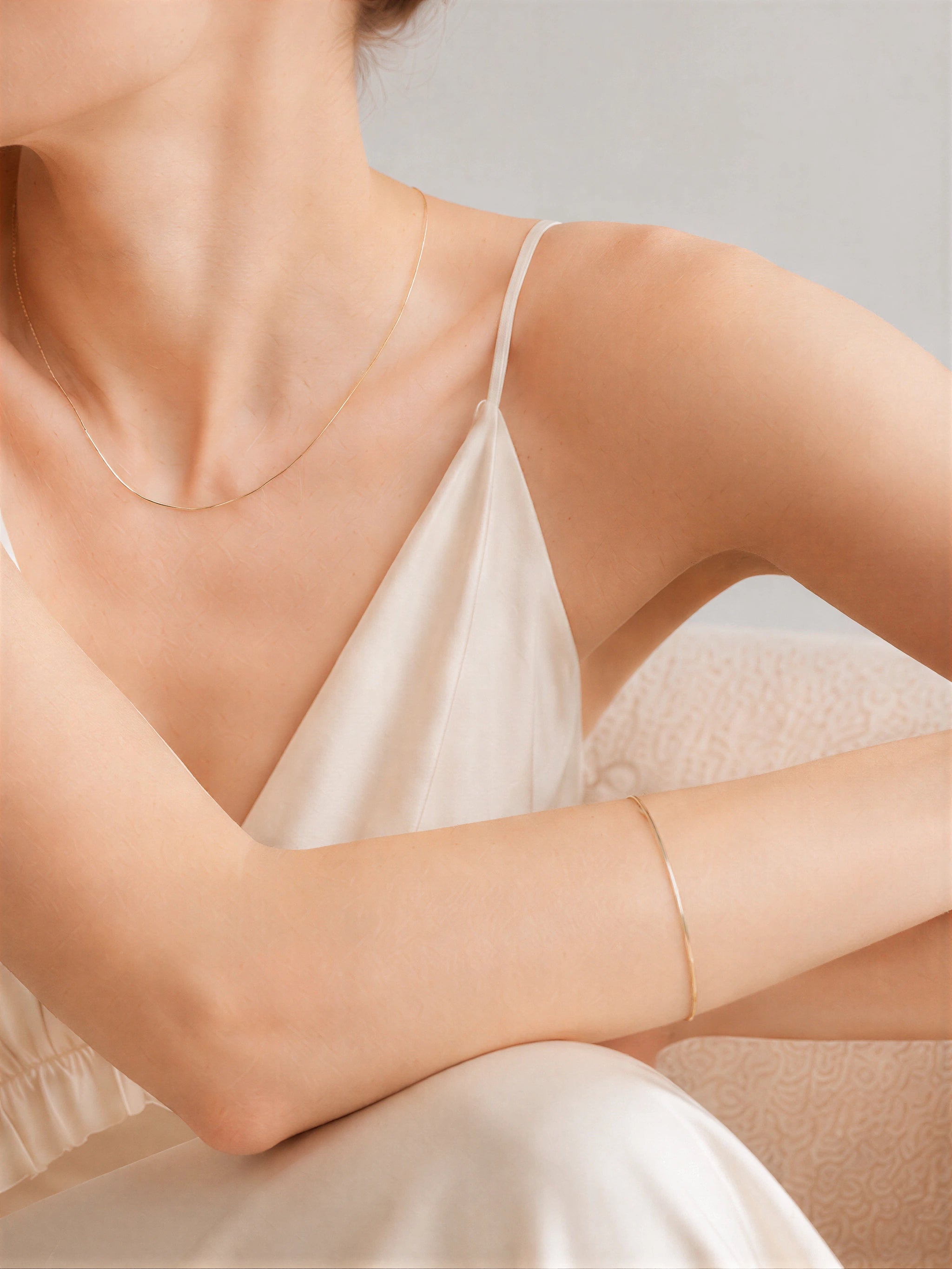 18K solid gold snake chain bracelet worn on wrist alongside dainty gold snake chain necklace, model seated in cream satin dress against neutral background