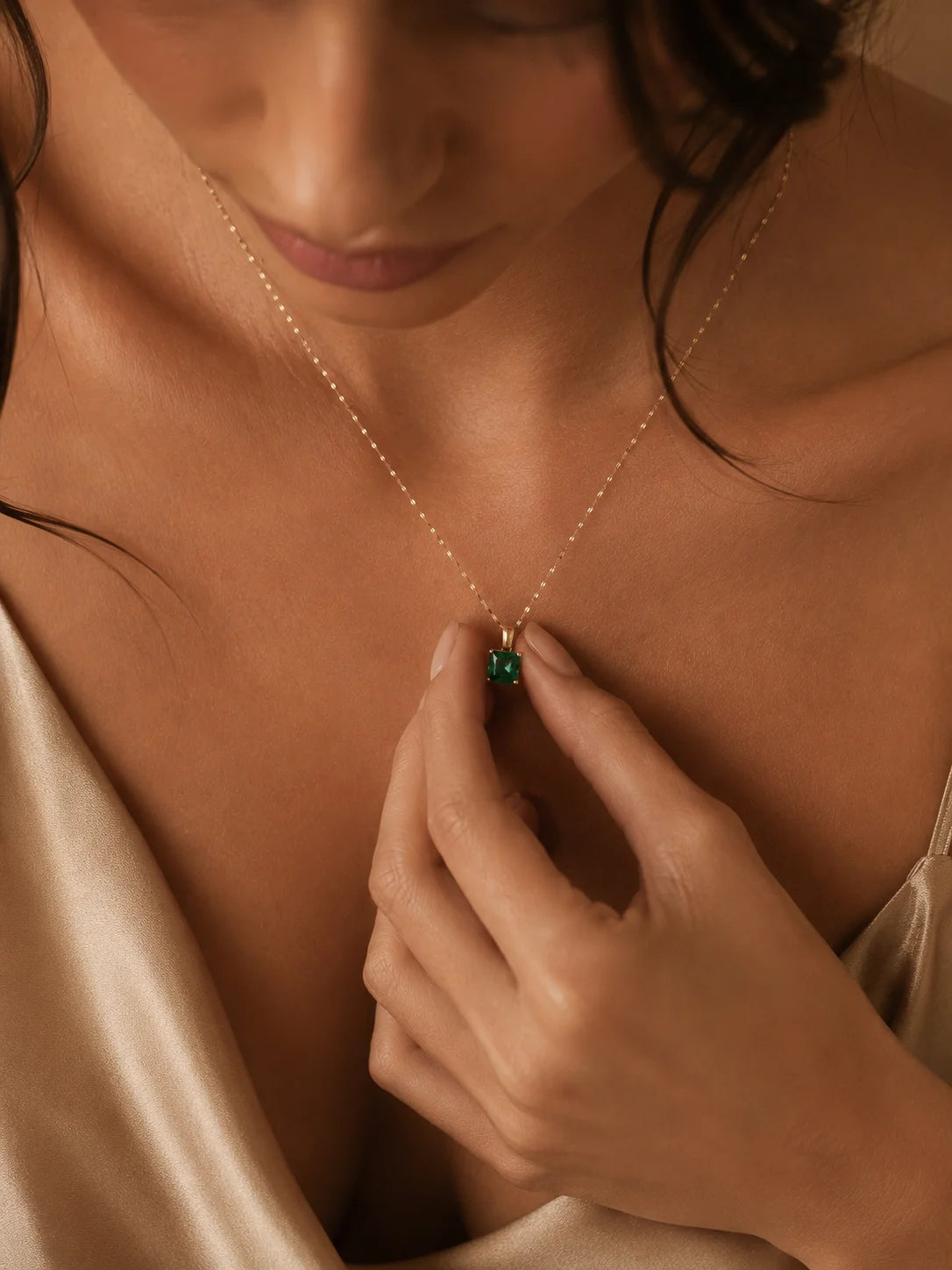A close-up lifestyle shot of a model wearing a delicate gold chain necklace with a small square-cut green gemstone pendant, wearing a champagne-colored satin top.