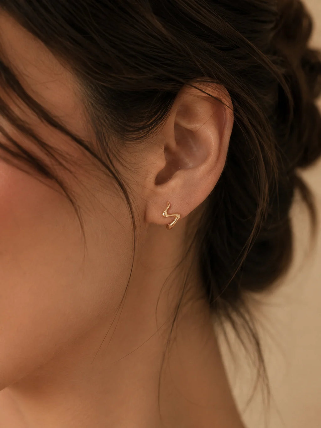 A close-up of a model's ear wearing a small, minimalist gold lightning bolt stud earring.