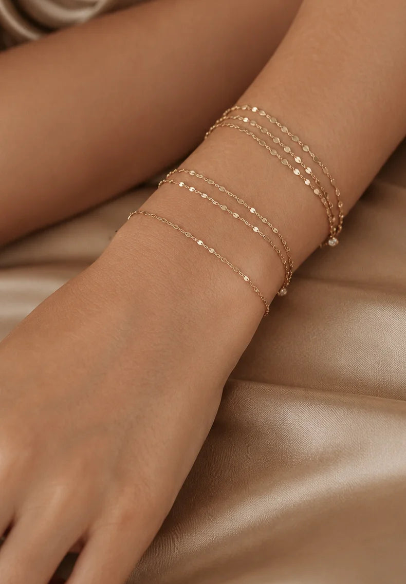 A close-up of a person's wrist and hand wearing multiple delicate rose gold chain bracelets, resting on a soft beige satin fabric.