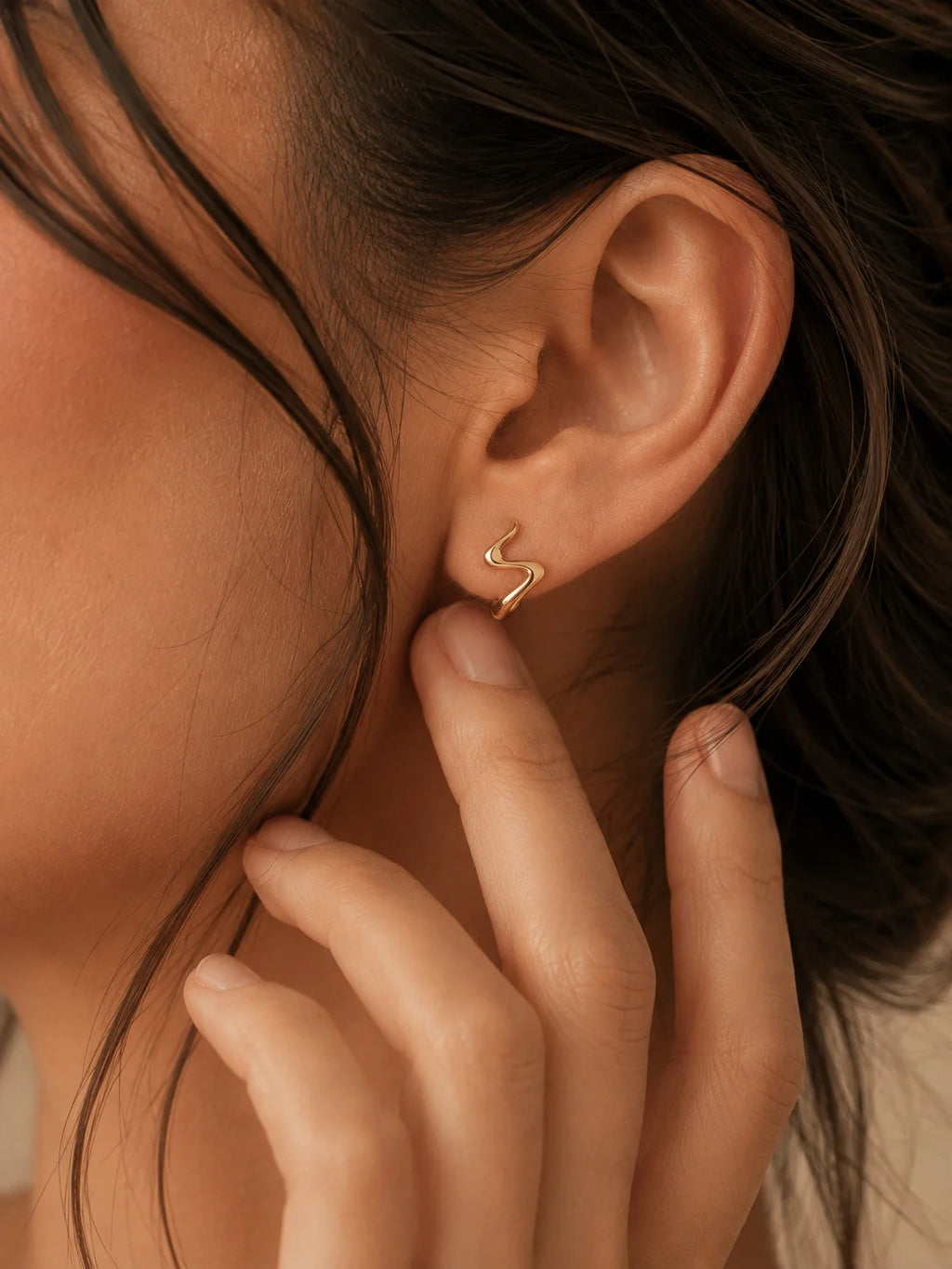 A close-up of a model's ear wearing a delicate, thin gold S-shaped stud earring, with her fingers gently touching her cheek.