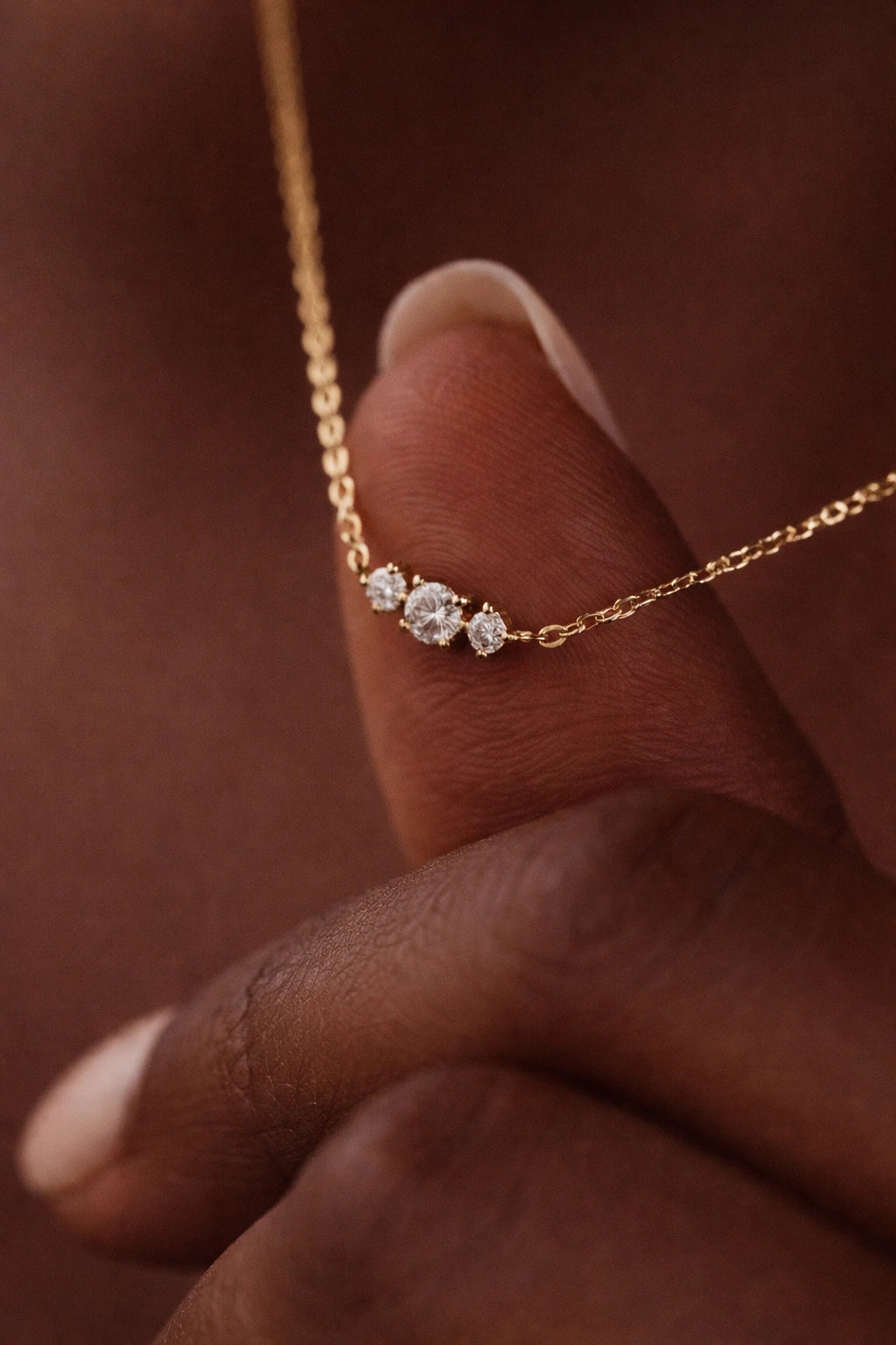 Gold necklace with three small diamonds held by a hand against a brown background