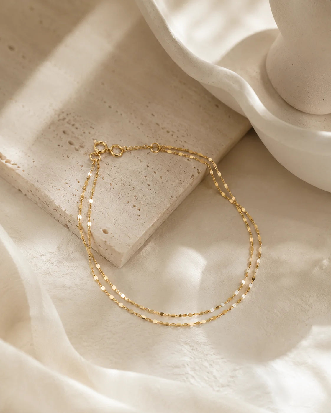 A delicate gold chain necklace with a small lobster clasp, draped over a textured beige stone slab and white fabric.