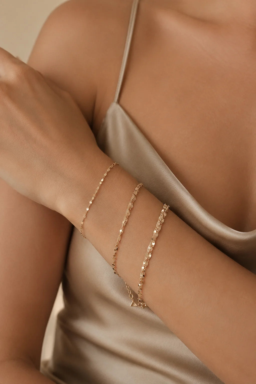 A close-up of a model's forearm wearing three delicate rose gold chain bracelets, paired with a satin champagne-colored camisole.